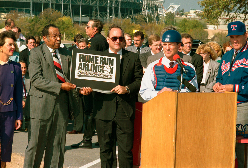 Arlington Mayor Richard Greene at podium speaking while Councilmember Elzie Odom helps to hold sign that reads "Home Run Arlington, Let's Keep The Rangers."