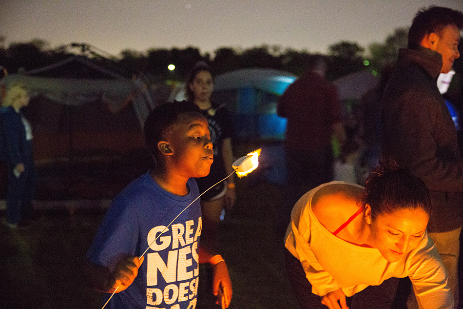 Boy holding a stick and blowing out fire from a fire roasted marshmallow. Woman nearby looking at fire.