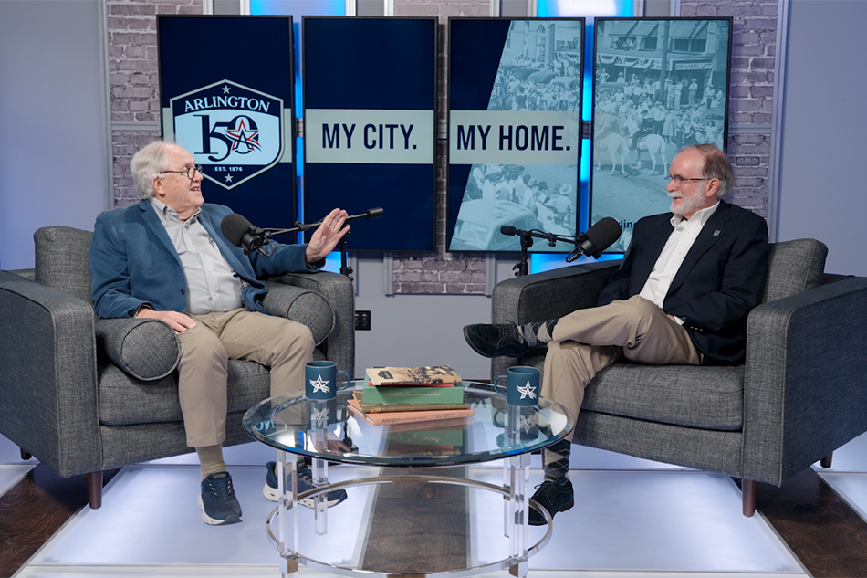 OK Carter, left, speaks as Gerald Saxon, right, laughs. Both are seated. Background image on TV screens say "Arlington 150. My City. My Home."