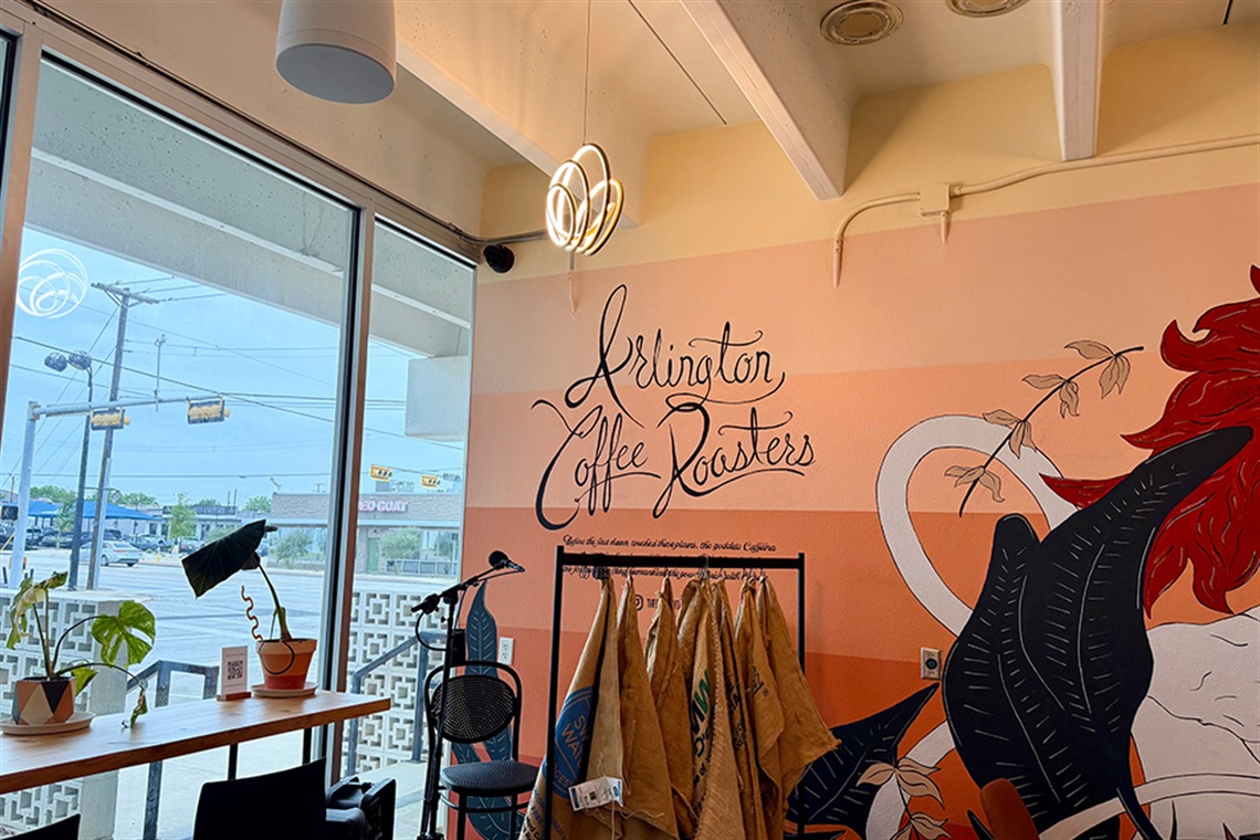 Colorful mural with the words "Arlington Coffee Roasters" painted on it, with a brightly lit window in front of a bar with plants set on top.
