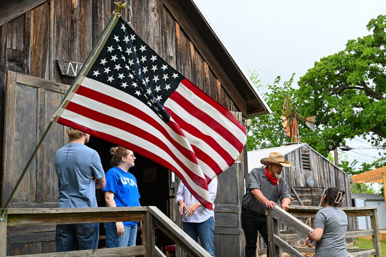 People talking with one another standing on the steps of a historic wooden schoolhouse.