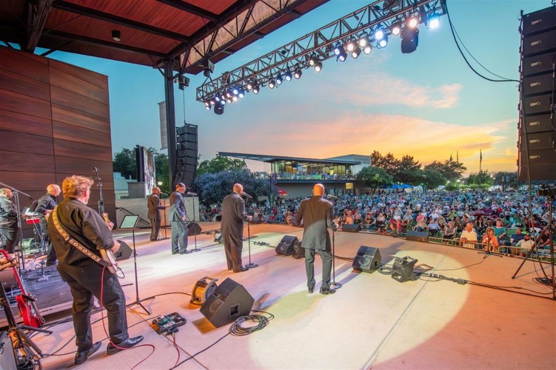 Band performing on stage at dusk with hundreds of people on the lawn enjoying the concert.