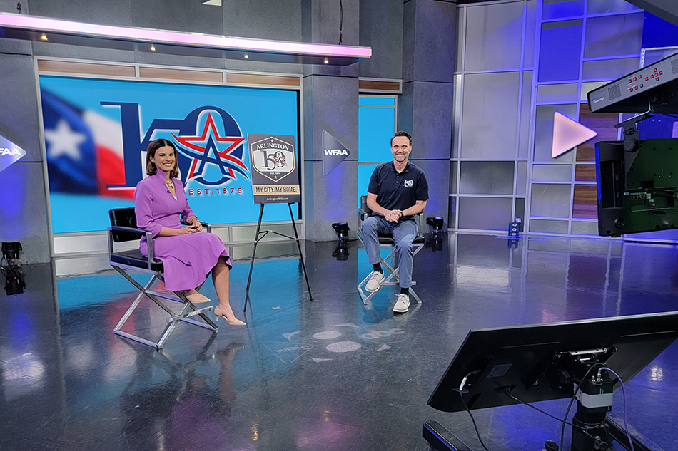 A TV studio setup featuring two people seated on tall chairs. The background displays a large logo with "WFAA" and "150 Years" along with a Texas flag motif. There's professional lighting, a camera, and a monitor visible in the studio.